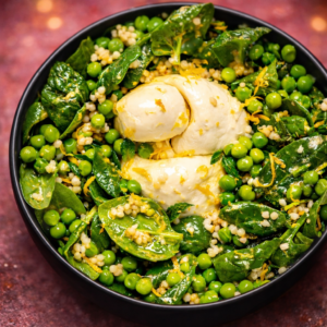 Bowl of spring-green pasta salad with baby spinach, sweet peas, fresh mint, and creamy burrata on top with a bright vinaigrette.