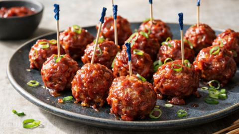 Glossy cranberry-chili glazed cocktail meatballs topped with sliced green onions and served with toothpicks on a dark plate.